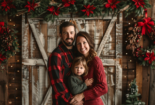 Family of three posing in front of a rustic wood barn door backdrop with red poinsettia garland.